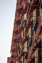 A tall building facade adorned with multiple red awnings and flower boxes mounted on the exterior. Each window has a triangular red canopy extending outward, providing shade and a decorative element. The flower boxes underneath are filled with small green plants.