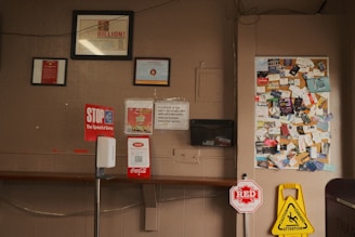 The image shows a bulletin board filled with various business cards and flyers. Below the board, there are several signs including a 'STOP the Spread of Germs' sign and a caution wet floor sign. There is also a hand sanitizer dispenser attached to a vertical stand in the foreground. The wall is a light brown color with some framed documents hanging above the bulletin board.