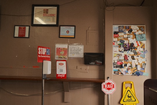 The image shows a bulletin board filled with various business cards and flyers. Below the board, there are several signs including a 'STOP the Spread of Germs' sign and a caution wet floor sign. There is also a hand sanitizer dispenser attached to a vertical stand in the foreground. The wall is a light brown color with some framed documents hanging above the bulletin board.