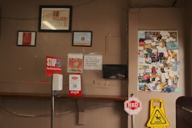 The image shows a bulletin board filled with various business cards and flyers. Below the board, there are several signs including a 'STOP the Spread of Germs' sign and a caution wet floor sign. There is also a hand sanitizer dispenser attached to a vertical stand in the foreground. The wall is a light brown color with some framed documents hanging above the bulletin board.
