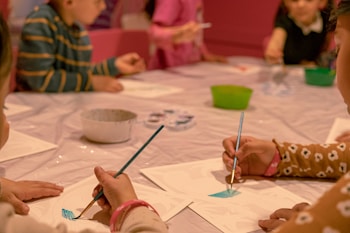 Children are seated around a table engaged in a painting activity. Each child holds a paintbrush, focusing on their artwork. A variety of colors and art supplies are scattered on the table, creating a lively and creative atmosphere.