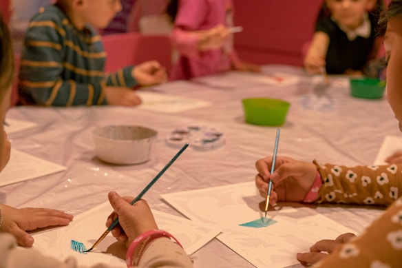 Children are seated around a table engaged in a painting activity. Each child holds a paintbrush, focusing on their artwork. A variety of colors and art supplies are scattered on the table, creating a lively and creative atmosphere.