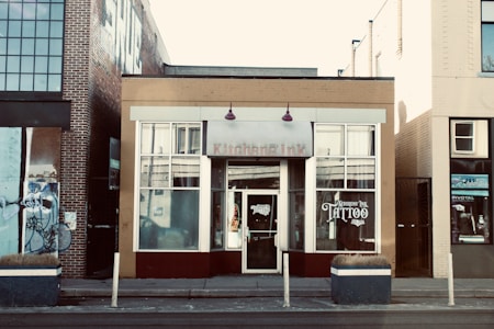 The scene features a street view of a tattoo parlor located in a small building among other urban structures. The central building has a beige and maroon facade, with large windows displaying the word 'TATTOO' prominently. A brick building is adjacent on the left side, adorned with graffiti artwork, while the building on the right has a yellow brick exterior. Small planters with dried plants are placed in front of the tattoo shop.