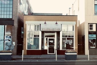 The scene features a street view of a tattoo parlor located in a small building among other urban structures. The central building has a beige and maroon facade, with large windows displaying the word 'TATTOO' prominently. A brick building is adjacent on the left side, adorned with graffiti artwork, while the building on the right has a yellow brick exterior. Small planters with dried plants are placed in front of the tattoo shop.