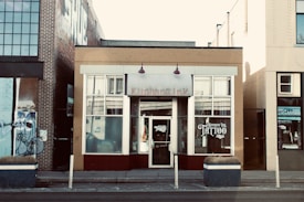The scene features a street view of a tattoo parlor located in a small building among other urban structures. The central building has a beige and maroon facade, with large windows displaying the word 'TATTOO' prominently. A brick building is adjacent on the left side, adorned with graffiti artwork, while the building on the right has a yellow brick exterior. Small planters with dried plants are placed in front of the tattoo shop.
