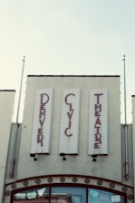 Vintage-style signage for the 'Denver Civic Theatre' in red lettering against a pale background. The building façade features decorative elements and two flagpoles, while banners or decorations are visible through the glass windows below.