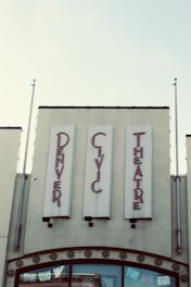 Vintage-style signage for the 'Denver Civic Theatre' in red lettering against a pale background. The building façade features decorative elements and two flagpoles, while banners or decorations are visible through the glass windows below.