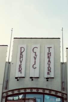 Vintage-style signage for the 'Denver Civic Theatre' in red lettering against a pale background. The building façade features decorative elements and two flagpoles, while banners or decorations are visible through the glass windows below.