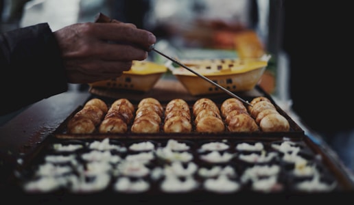 A hand is using a skewer to cook and turn several round, golden-brown snacks in a grill pan. In the foreground, chopped onions are arranged on the cooking surface. The background contains blurred yellow containers, indicating a food preparation setting.