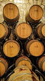 Wooden barrels are stacked against a stone wall. They have a circular design and are held in black metal racks. The surface of the barrels displays stamped logos or graphics.