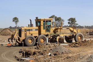A large yellow construction grader sits on a dirt construction site, surrounded by piles of earth. It is clearly marked with the brand 'Caterpillar' and features six large wheels. In the background, there are a few scattered trees and a clear blue sky.