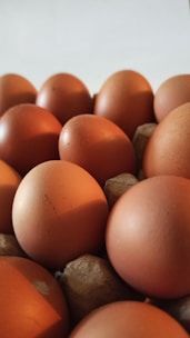 A close-up of several brown eggs arranged in a cardboard egg carton. The eggs are tightly packed and have a smooth texture, with some displaying small blemishes or imperfections on their surface.