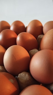 A close-up of several brown eggs arranged in a cardboard egg carton. The eggs are tightly packed and have a smooth texture, with some displaying small blemishes or imperfections on their surface.