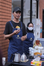 A person wearing a black hat and a blue apron is holding a small cup, standing at an outdoor setup with brick walls in the background. Next to them, another person in a blue apron and a mask stands, appearing to be assisting or observing. On the table in front of them are various coffee-making tools including a kettle, a grinder, and cups. The apron has 'Lestari Coffee' printed on it.