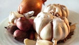 A close-up of a white plate holding various types of onions and garlic. Large bulbs of garlic and onions are prominently placed along with smaller shallots. The background is plain and out of focus, highlighting the vegetables.