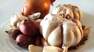 A close-up of a white plate holding various types of onions and garlic. Large bulbs of garlic and onions are prominently placed along with smaller shallots. The background is plain and out of focus, highlighting the vegetables.