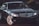 A sleek black Mercedes-Benz sedan is parked on a street, adorned with glowing headlights and reflecting the branches of nearby trees on its polished surface. The emblem is prominently displayed on the grille, and the car's design emphasizes luxury and elegance.