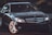 A sleek black Mercedes-Benz sedan is parked on a street, adorned with glowing headlights and reflecting the branches of nearby trees on its polished surface. The emblem is prominently displayed on the grille, and the car's design emphasizes luxury and elegance.