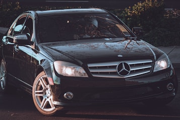 A sleek black Mercedes-Benz sedan is parked on a street, adorned with glowing headlights and reflecting the branches of nearby trees on its polished surface. The emblem is prominently displayed on the grille, and the car's design emphasizes luxury and elegance.