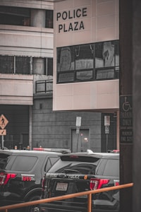 Several black police vehicles are parked outside a building labeled Police Plaza. The area appears urban, with modern architectural elements and tall structures in the background. There are signs indicating emergency services and parking restrictions.