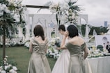 A wedding scene outdoors with three women, one wearing a white dress and holding a bouquet, possibly the bride. The other two are in matching olive-colored dresses. The background is decorated with white and green floral arrangements under a cloudy sky. A table is set with drinks and a framed photo.