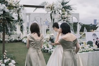 A wedding scene outdoors with three women, one wearing a white dress and holding a bouquet, possibly the bride. The other two are in matching olive-colored dresses. The background is decorated with white and green floral arrangements under a cloudy sky. A table is set with drinks and a framed photo.