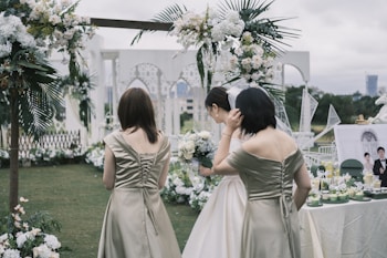 A wedding scene outdoors with three women, one wearing a white dress and holding a bouquet, possibly the bride. The other two are in matching olive-colored dresses. The background is decorated with white and green floral arrangements under a cloudy sky. A table is set with drinks and a framed photo.