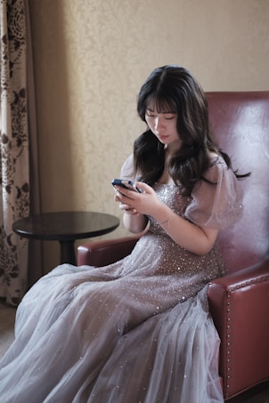 A woman in a glamorous, sparkling gown sits in a plush red armchair, intently focused on a smartphone. The background features an ornamental curtain and a wall with decorative patterns.