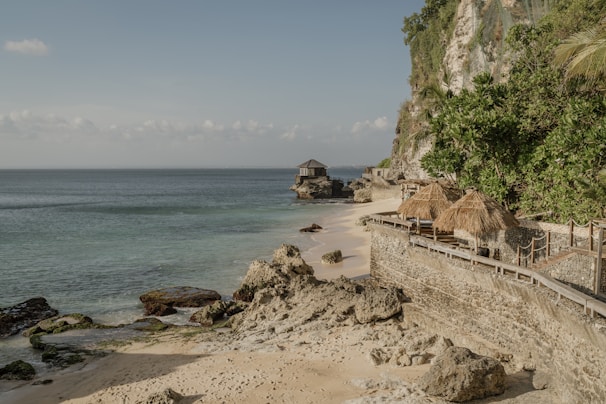 A serene coastal scene features a sandy beach bordered by rocky outcrops and a lush, green cliffside. Traditional straw-roofed cabanas line a stone pathway that curves alongside the water. The sea is calm and stretches out to the horizon under a soft blue sky with a few clouds.