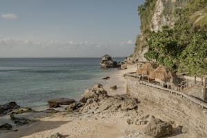 A serene coastal scene features a sandy beach bordered by rocky outcrops and a lush, green cliffside. Traditional straw-roofed cabanas line a stone pathway that curves alongside the water. The sea is calm and stretches out to the horizon under a soft blue sky with a few clouds.