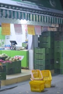 A small market stall displays various fruits and vegetables in green plastic crates. Yellow shopping baskets are stacked in the foreground. Overhead, signs in both English and another language advertise deals of the day. A striped green and white awning partially shades the entrance, and a cashier's counter with an electronic register is visible inside.