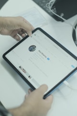 A person is holding a tablet that displays a social media profile on its screen. The individual's profile shows a profile picture and various profile details. The tablet is connected to a charging cable. The background includes a laptop and some papers on the desk.