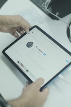 A person is holding a tablet that displays a social media profile on its screen. The individual's profile shows a profile picture and various profile details. The tablet is connected to a charging cable. The background includes a laptop and some papers on the desk.