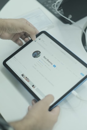 A person is holding a tablet that displays a social media profile on its screen. The individual's profile shows a profile picture and various profile details. The tablet is connected to a charging cable. The background includes a laptop and some papers on the desk.