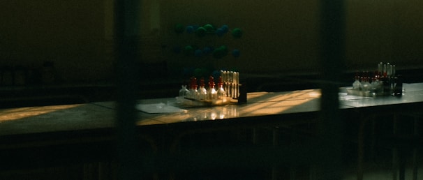 A dimly lit laboratory table with an arrangement of test tubes, some filled with liquid in varied colors. There is also a molecular model in the background. Sunlight through the window casts dramatic shadows and highlights on the surface.