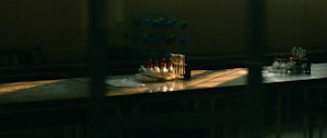 A dimly lit laboratory table with an arrangement of test tubes, some filled with liquid in varied colors. There is also a molecular model in the background. Sunlight through the window casts dramatic shadows and highlights on the surface.