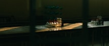 A dimly lit laboratory table with an arrangement of test tubes, some filled with liquid in varied colors. There is also a molecular model in the background. Sunlight through the window casts dramatic shadows and highlights on the surface.