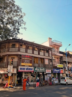 A busy street scene featuring an old two-story building with various businesses on the ground floor, including a bakery, a grocery store, and a dairy shop. The structure has intricate balcony railings and signs in a local language. People are walking and engaging in daily activities; one person is dressed in orange attire and stands out prominently. There is also a tree to the left, and a blue sky visible overhead.
