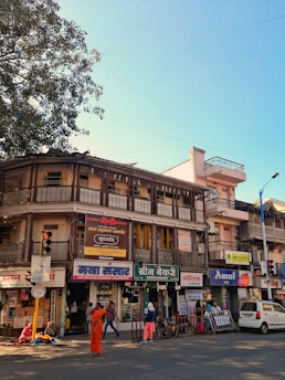 A busy street scene featuring an old two-story building with various businesses on the ground floor, including a bakery, a grocery store, and a dairy shop. The structure has intricate balcony railings and signs in a local language. People are walking and engaging in daily activities; one person is dressed in orange attire and stands out prominently. There is also a tree to the left, and a blue sky visible overhead.