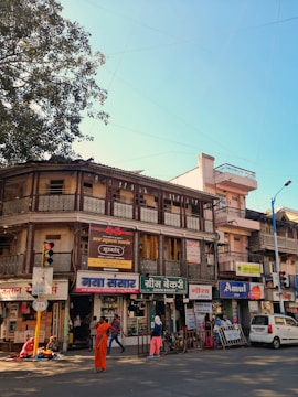 A busy street scene featuring an old two-story building with various businesses on the ground floor, including a bakery, a grocery store, and a dairy shop. The structure has intricate balcony railings and signs in a local language. People are walking and engaging in daily activities; one person is dressed in orange attire and stands out prominently. There is also a tree to the left, and a blue sky visible overhead.