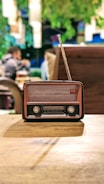 An antique radio with a brown and black casing, positioned on a wooden table. In the background, blurred figures of people and greenery provide a cozy, vintage ambiance.