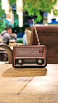 An antique radio with a brown and black casing, positioned on a wooden table. In the background, blurred figures of people and greenery provide a cozy, vintage ambiance.