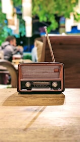 An antique radio with a brown and black casing, positioned on a wooden table. In the background, blurred figures of people and greenery provide a cozy, vintage ambiance.