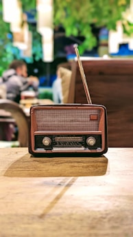 An antique radio with a brown and black casing, positioned on a wooden table. In the background, blurred figures of people and greenery provide a cozy, vintage ambiance.