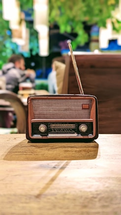 An antique radio with a brown and black casing, positioned on a wooden table. In the background, blurred figures of people and greenery provide a cozy, vintage ambiance.