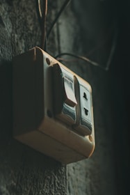A vintage-looking electrical switch with two buttons mounted on a textured wall. The switch is slightly dusty, suggesting it is old or used in an outdoor or rustic environment. The lighting is dim, casting shadows and giving the scene a moody atmosphere.