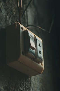 A vintage-looking electrical switch with two buttons mounted on a textured wall. The switch is slightly dusty, suggesting it is old or used in an outdoor or rustic environment. The lighting is dim, casting shadows and giving the scene a moody atmosphere.
