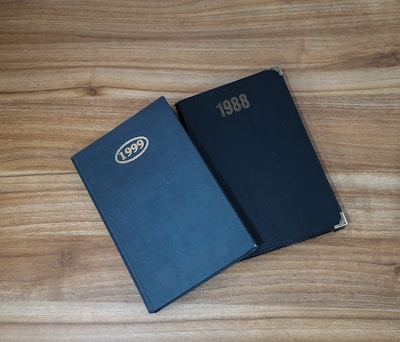 Two hardcover books are placed on a wooden surface. The book on the left is labeled 1999, and the one on the right is labeled 1988. Both books have a simple, solid black cover with gold embossed years.