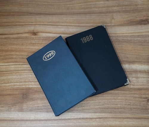 Two hardcover books are placed on a wooden surface. The book on the left is labeled 1999, and the one on the right is labeled 1988. Both books have a simple, solid black cover with gold embossed years.