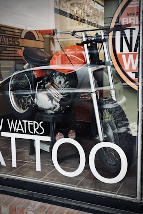 A motorcycle is displayed inside a shop with a large window. The bike features a bold orange fuel tank and is equipped with knobby tires, indicating off-road capabilities. Reflections on the glass suggest a street setting, and visible signage on the window includes the word 'TATTOO' in large white letters.
